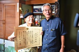 座喜味市長（右）から亀濵さんに祝い状が手渡された＝16日、平良西里