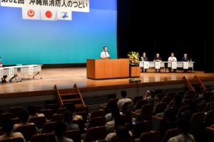 県消防人のつどいであいさつする照屋会長（壇上）＝26日、マティダ市民劇場