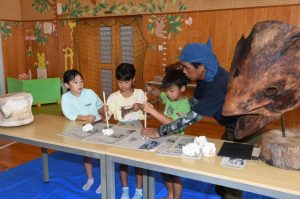 石の塊の中から化石を掘り出す子供たちと見守る平良さん（右）＝２５日、ひらら児童館