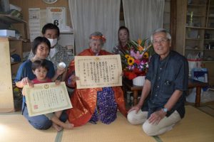 本村さん（中央）に内閣総理大臣から贈られた祝い状と銀杯を手渡した座喜味市長（右）と家族＝６日、市城辺福里の本村さんの自宅